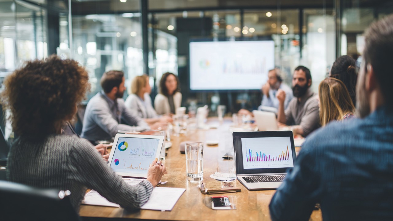 Diverse business team collaborating around a conference table reviewing digital tablets and laptops with user satisfaction graphs and website performance metrics in a modern, well-lit office.