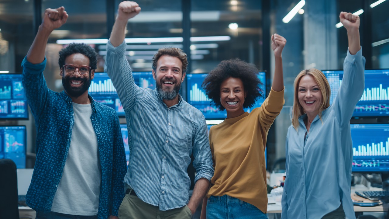 Diverse team celebrating success in a modern office with performance charts on screens, highlighting business growth and synthetic monitoring benefits.