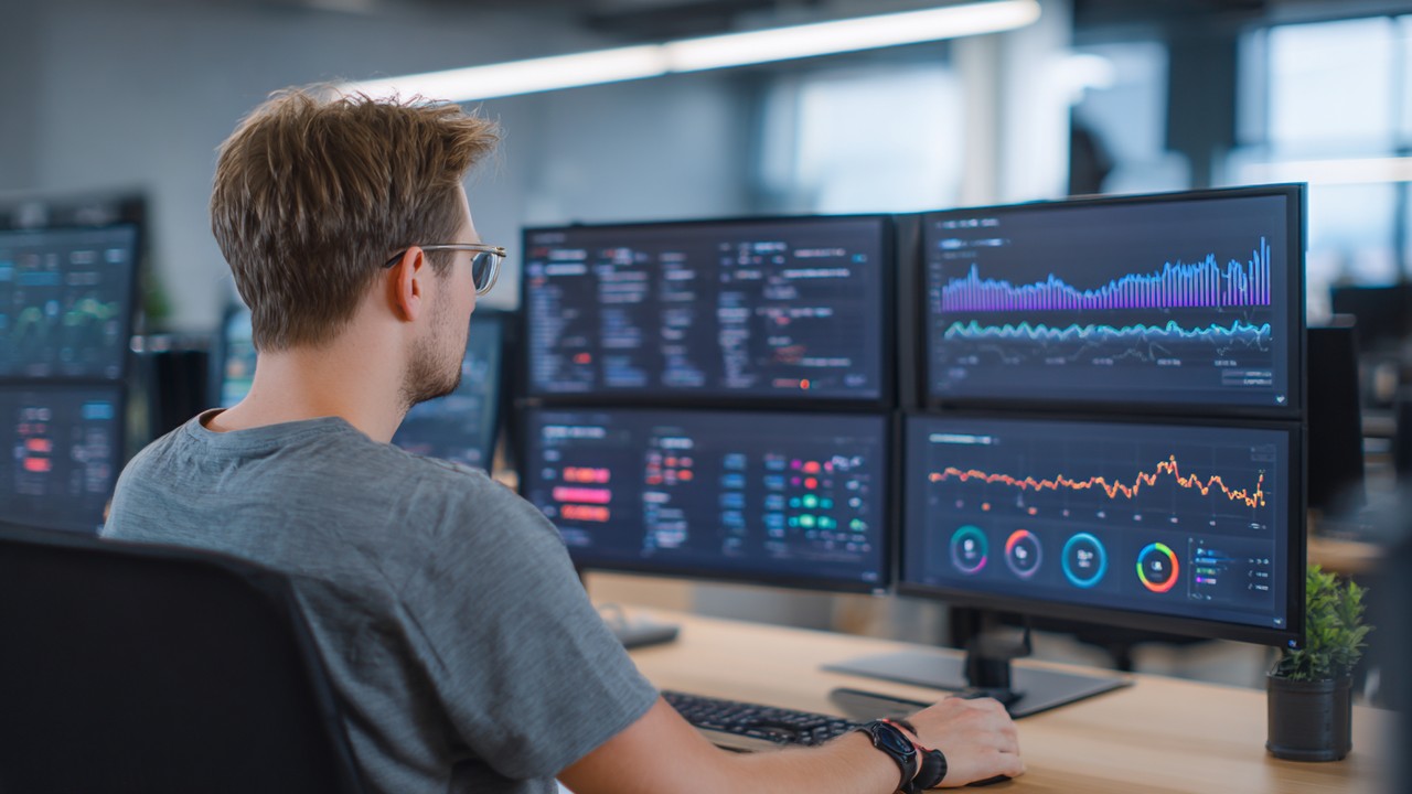 Modern web developer analyzing website performance metrics on multiple screens with graphs and server response times in a bright office.