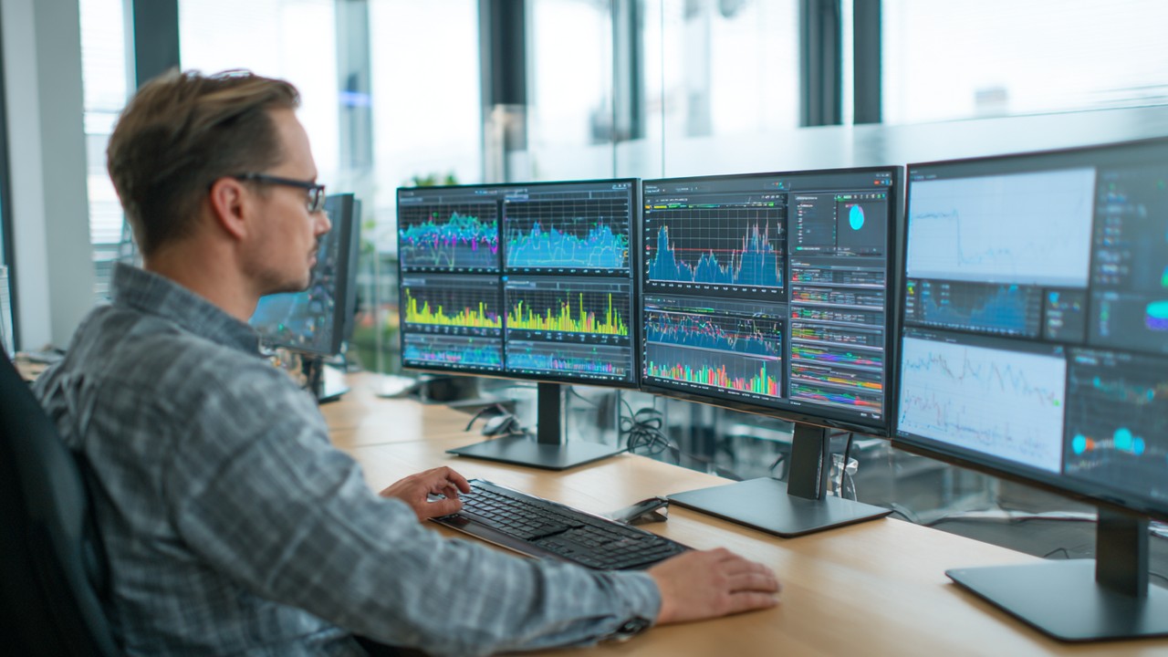 Professional analyzing website performance data on multiple monitors, displaying graphs and server response times in a bright office.