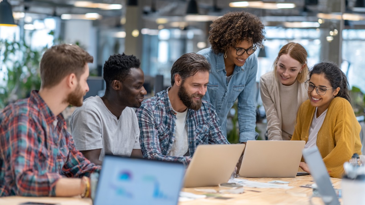 Diverse software developers collaborating around a table with laptops and tablets, reviewing performance metrics in a modern office.