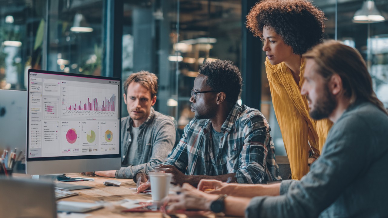 Diverse team of web developers, UX designers, and site managers collaborating around a computer displaying complex data charts in a modern office.