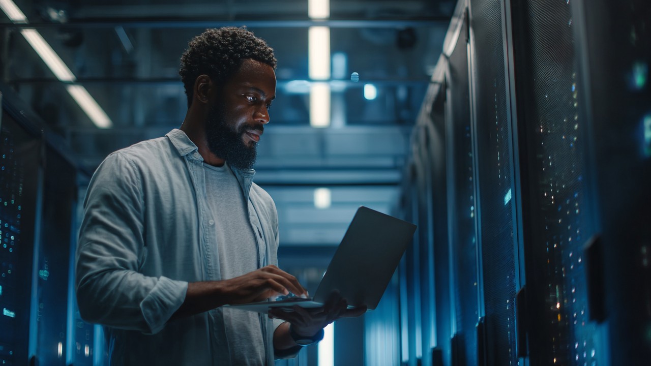 Modern data center server room with IT professional adjusting network settings on laptop, showcasing high-tech environment for traffic management and load balancing.