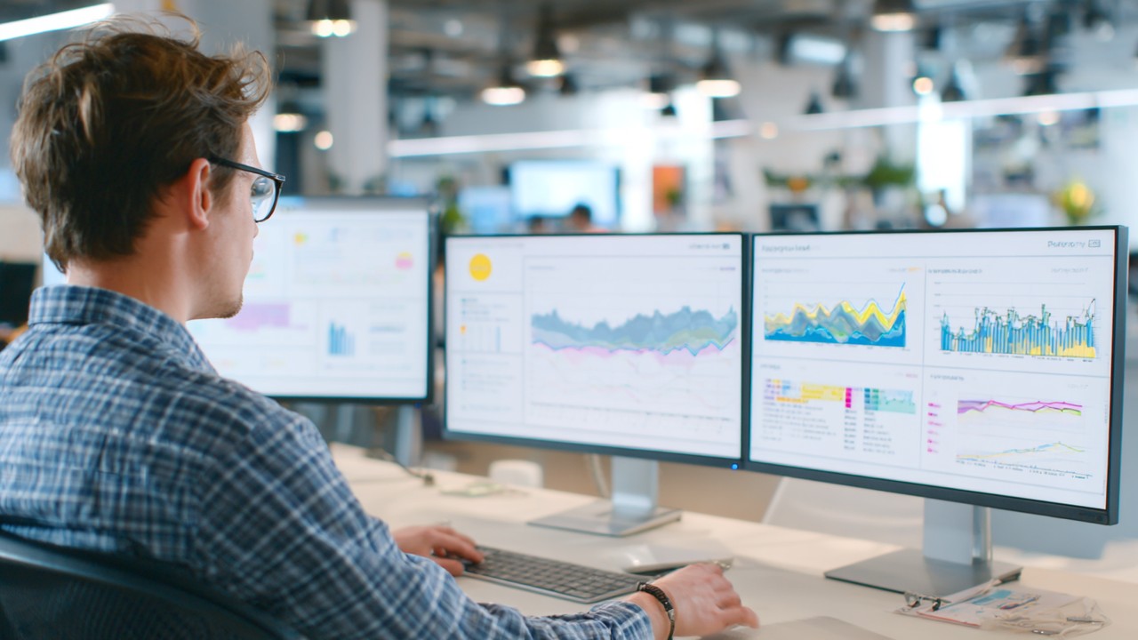 Person analyzing website performance data on multiple computer screens with graphs and charts in a modern office, emphasizing web monitoring and optimization.